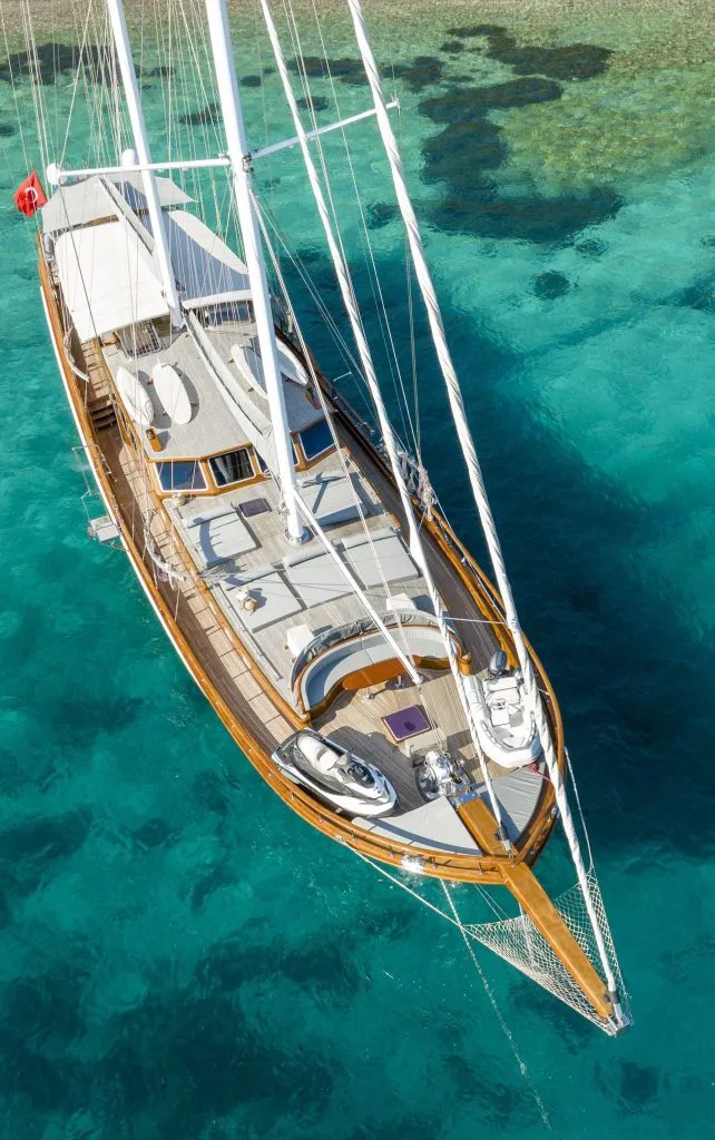 Aerial view of a large sailboat with wooden decking floating on clear turquoise water near a coral reef.