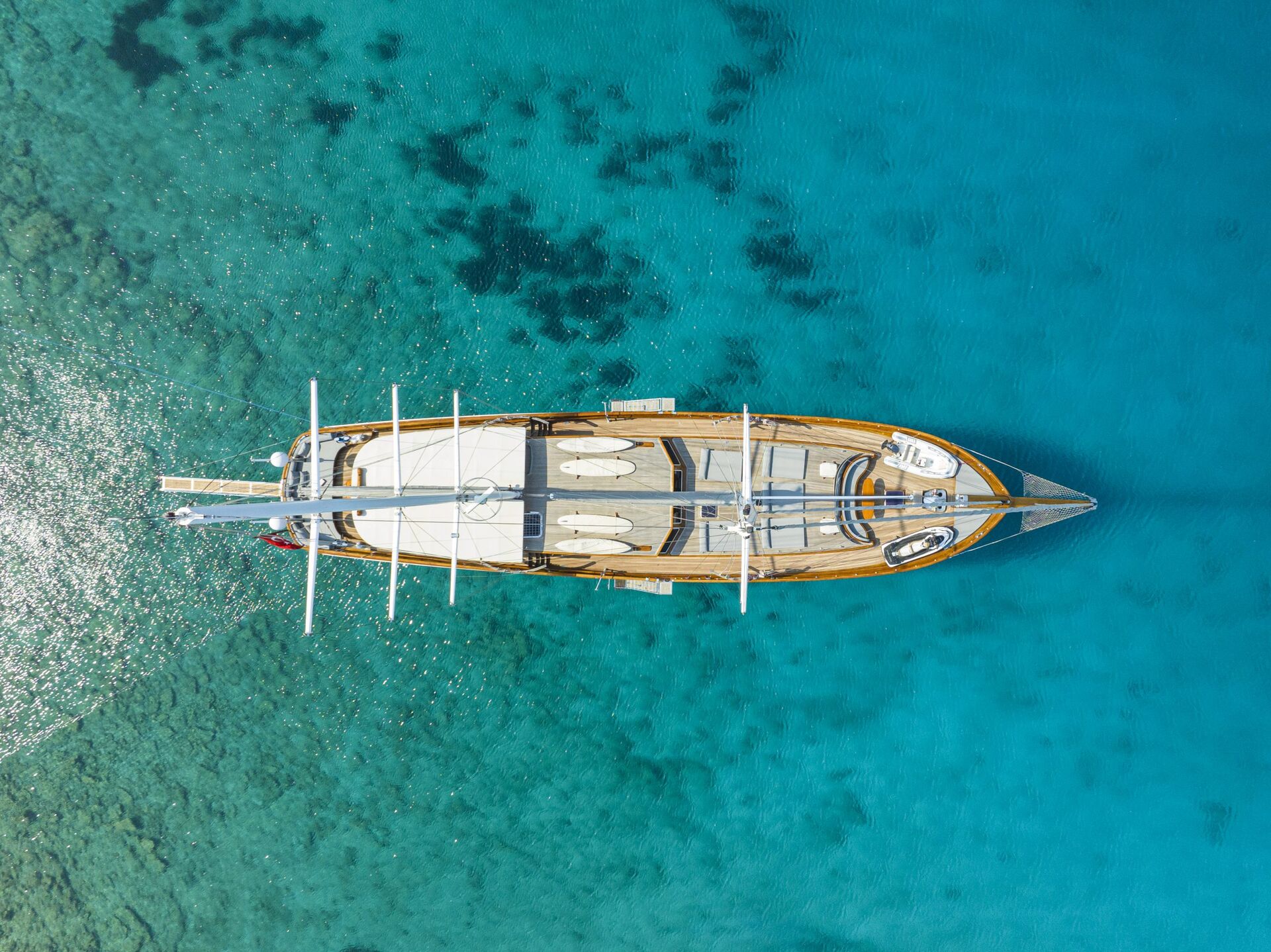 Aerial view of a large wooden sailboat anchored in clear, turquoise water. The boat is surrounded by calm sea, revealing the seabed below and creating a serene, picturesque scene.