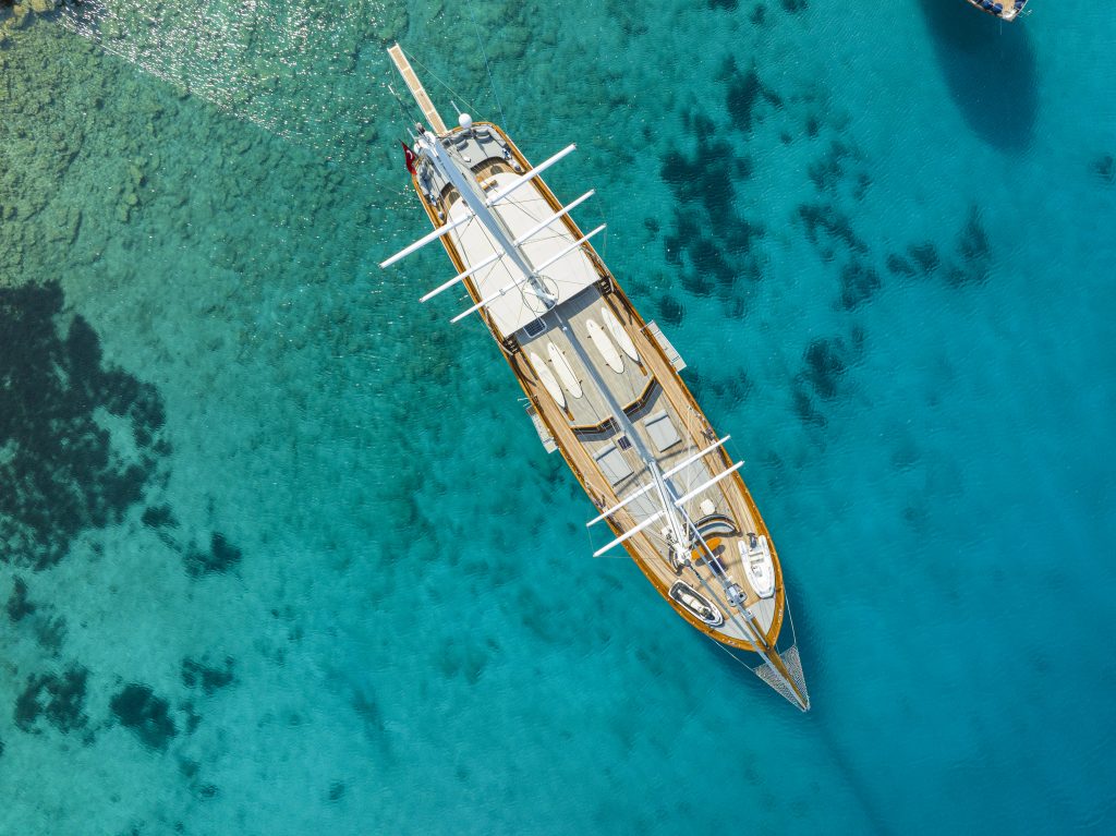 Aerial view of a large wooden sailboat with white sails, floating on clear turquoise water above a shallow seabed, casting a shadow below.