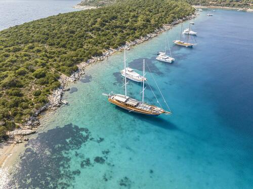 Aerial view of several sailboats and yachts anchored in clear turquoise water near a rocky, green coastline with dense vegetation. The boats are close to a small, sandy beach along the shore.