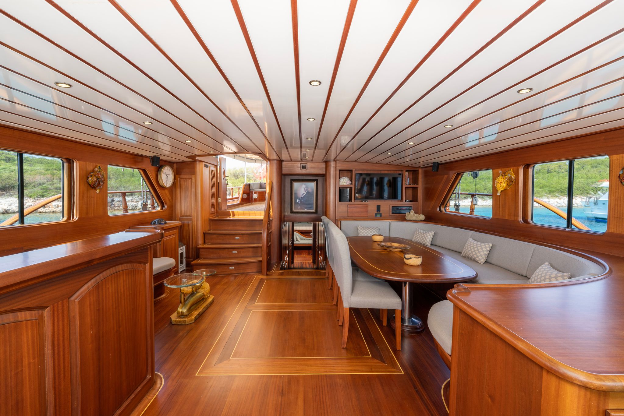Spacious yacht interior with polished wooden floors, walls, and ceiling, featuring a curved seating area with gray cushions, a dining table, bar counter, and large windows offering views of water and greenery.