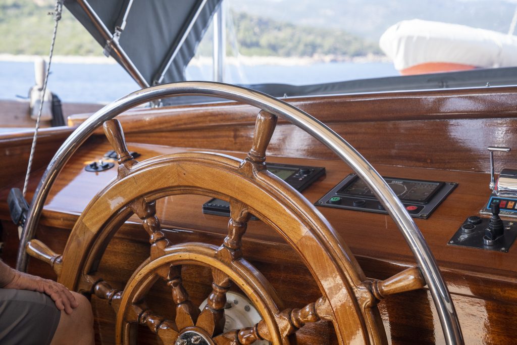 A close-up of a polished wooden ship’s steering wheel with metal accents, control panels, and navigation instruments on the wooden dashboard. A person’s arm is visible to the left, and water and coastline are seen in the background.