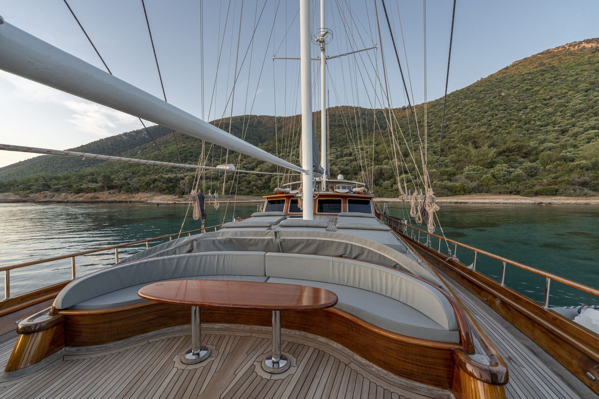 View from the deck of a luxury sailboat with a curved seating area and table, overlooking calm water and tree-covered hills under a clear sky.