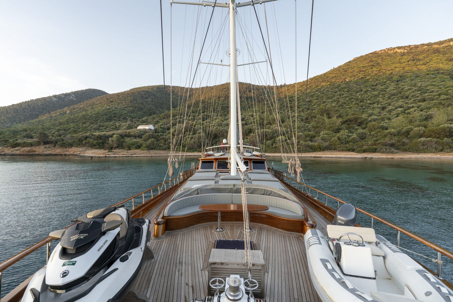 View from the deck of a yacht with jet skis and a small boat, anchored near a calm shoreline with green hills and trees under a clear sky.