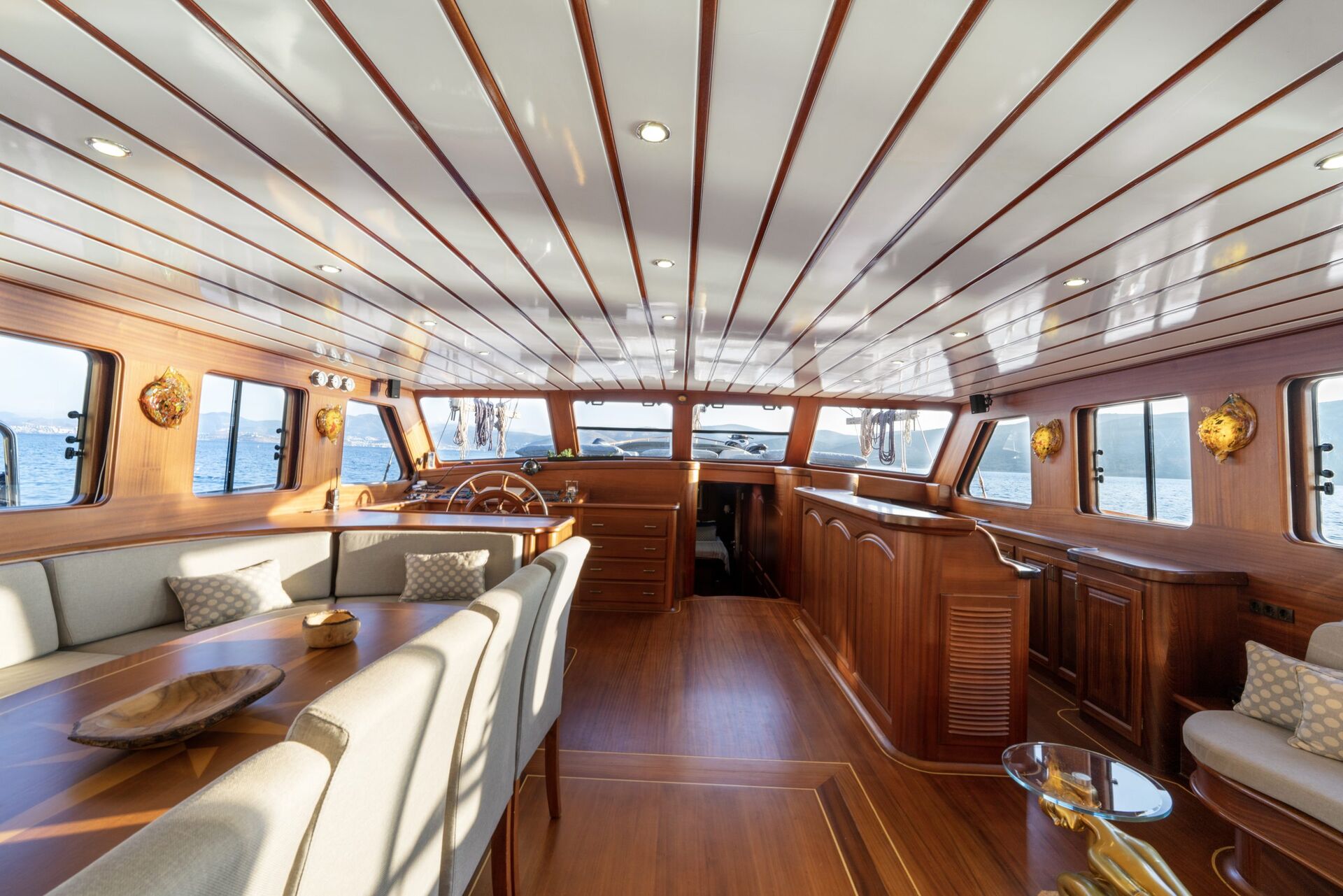 Spacious yacht interior with polished wood paneling, cream-colored sofas, a wooden dining table, and large windows offering ocean views; the captain’s wheel is visible near the front.