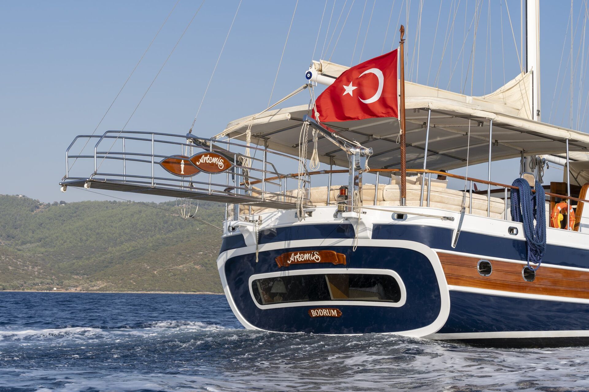 A large yacht with a Turkish flag sails on the sea near a green, hilly coastline. The yacht has wooden and white details, with the name “Artemis” displayed on the stern.