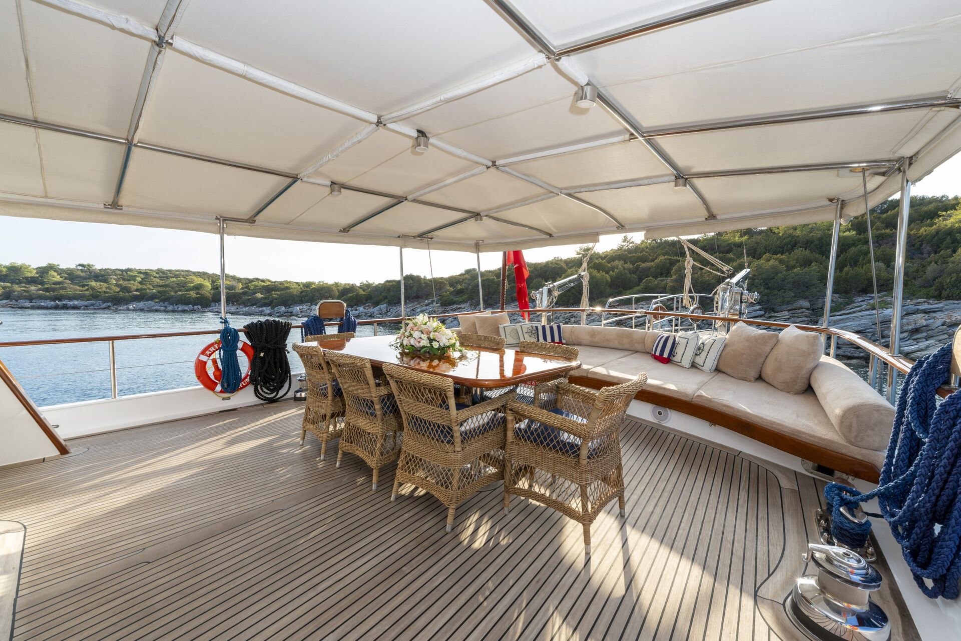 A spacious yacht deck with a covered seating area, wicker chairs around a wooden table, beige cushions on a curved bench, and scenic views of calm water and green shoreline in the background.