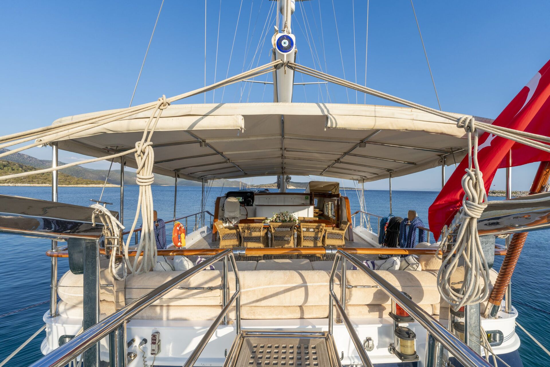 View from the rear of a yacht showing a shaded outdoor dining area with a wooden table and chairs, surrounded by railings, overlooking calm blue water and a clear sky.