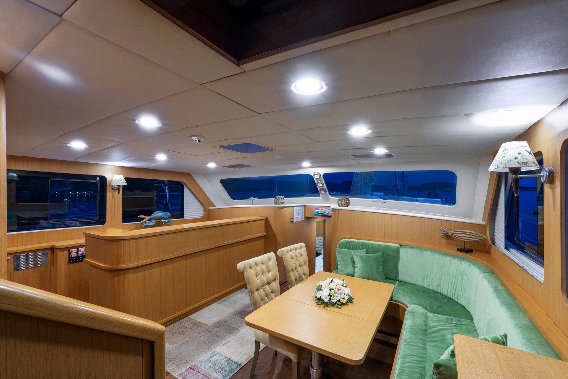 A cozy yacht interior with wood paneling, green cushioned seating, a wooden table with a flower arrangement, soft overhead lighting, and large windows showing a view of the harbor at dusk.