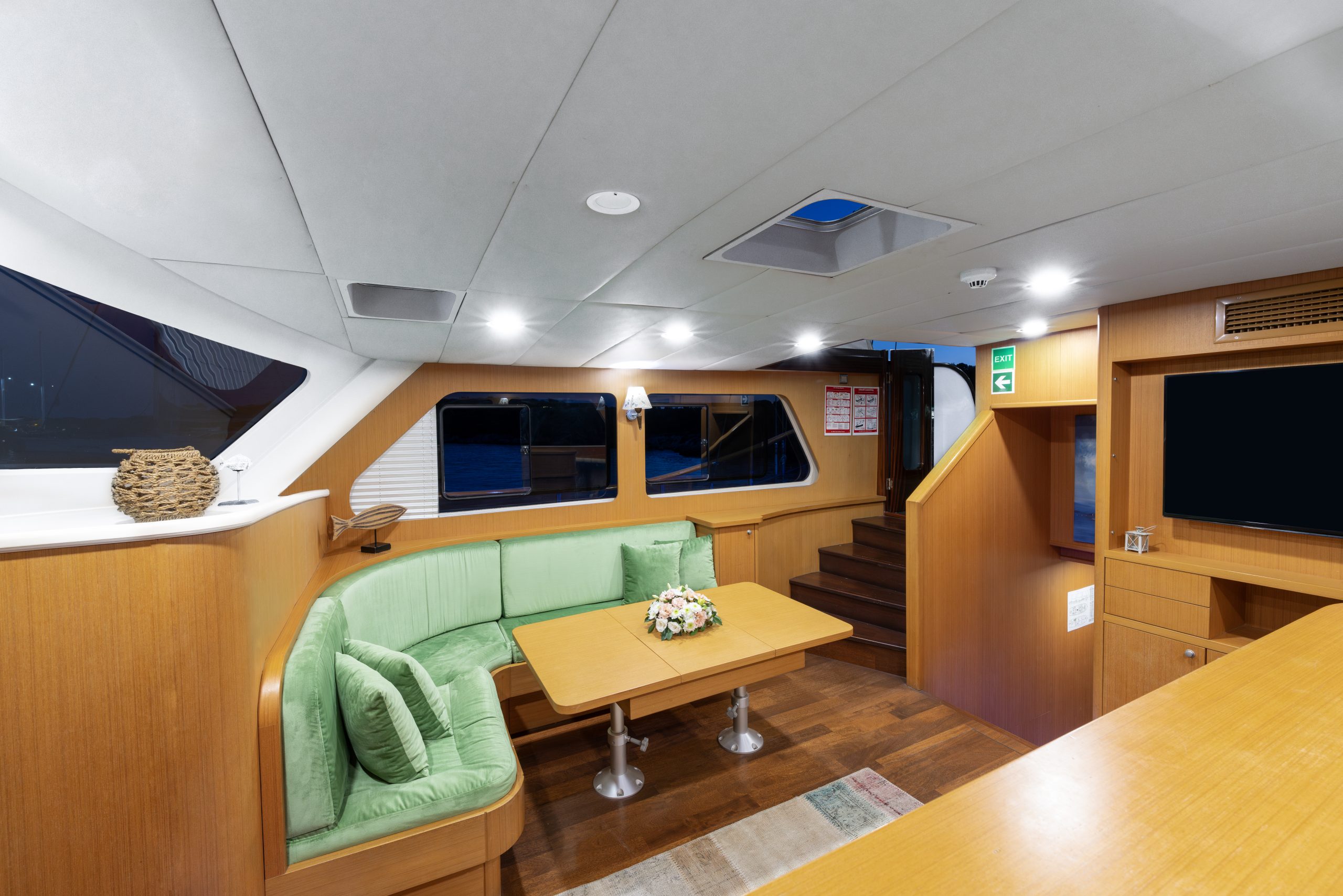 Modern yacht interior featuring a cozy green cushioned seating area around a wooden table, wood paneled walls, large windows, stairs leading up, and a flat-screen TV on the wall. Bright overhead lighting.