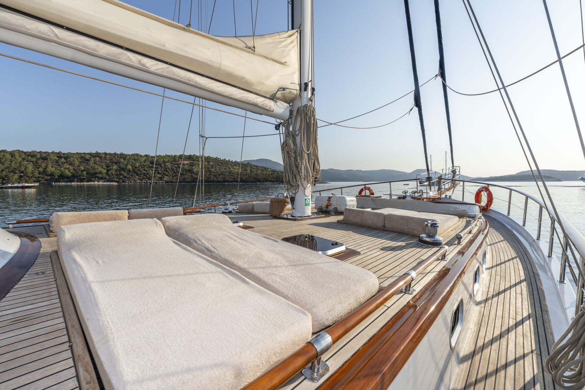 View from the deck of a yacht with cushioned sunbeds and polished wooden railings, overlooking calm water and a tree-lined shoreline under a clear sky.