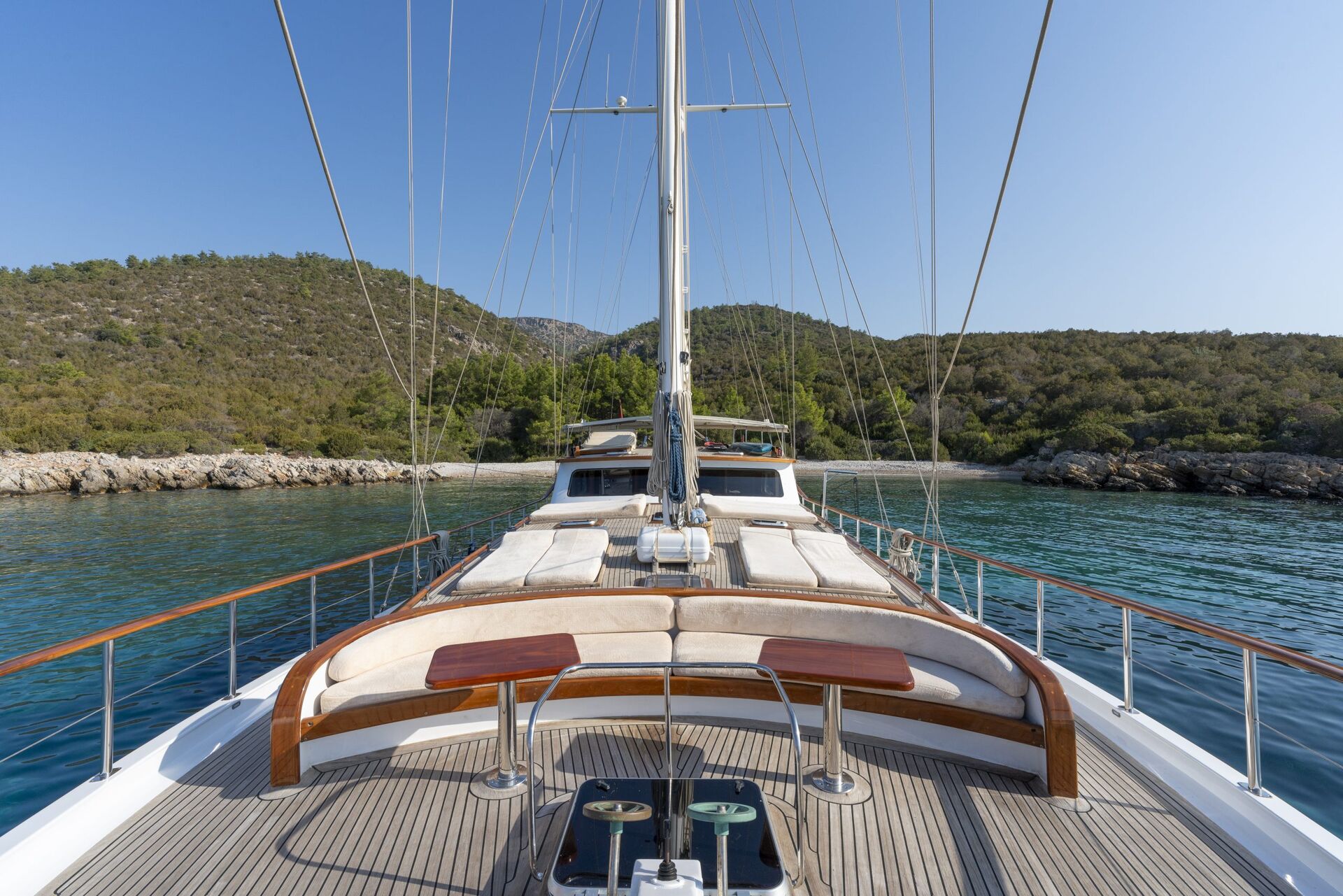 View from the deck of a yacht anchored near a rocky shoreline with green hills and trees in the background under a clear blue sky. The yacht has cushioned seating and small tables on the deck.