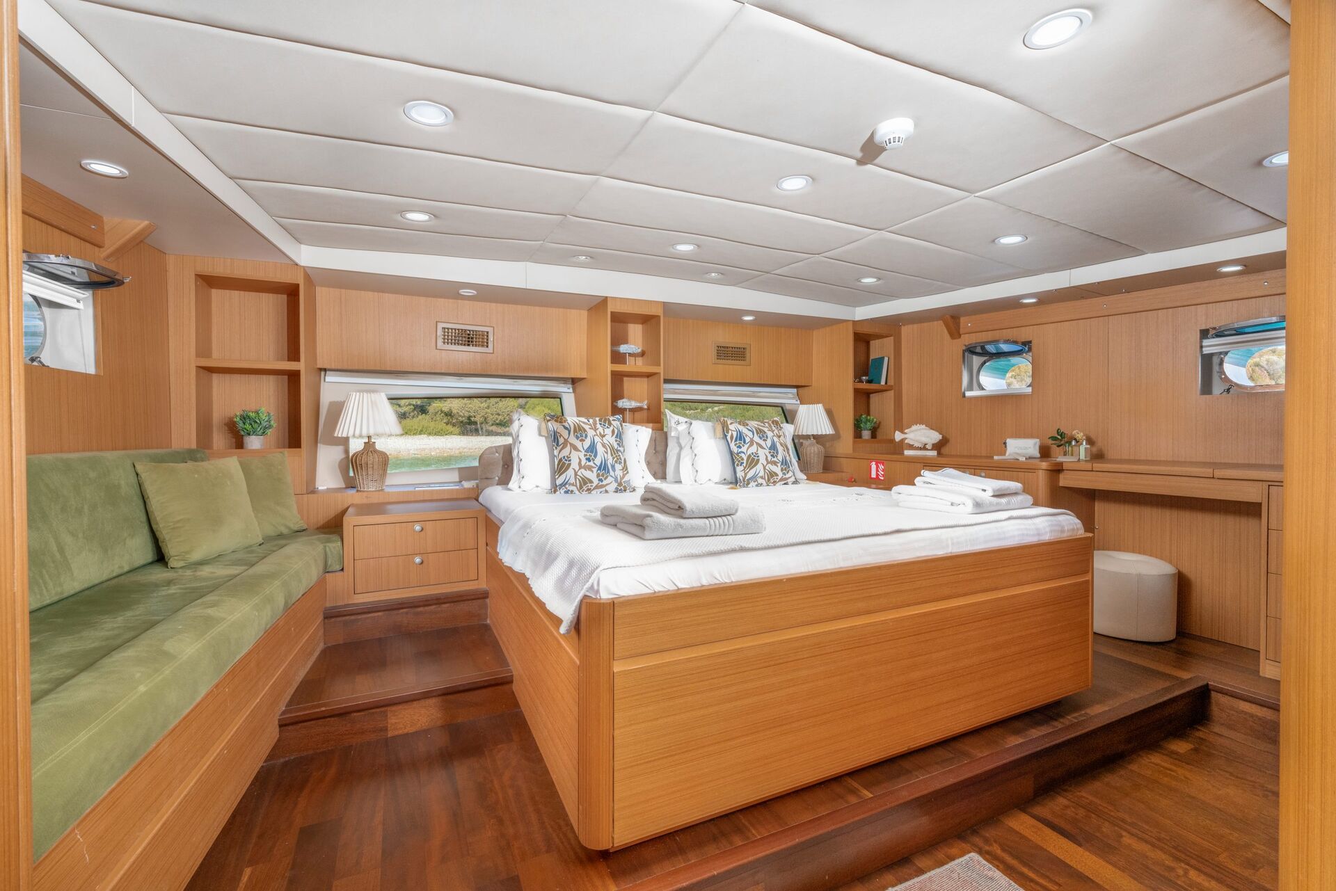 A spacious yacht cabin with a large bed, green sofa, wooden furniture, and decorative pillows. There are windows on the sides, built-in shelves with plants, and a small desk with a stool; the room looks bright and inviting.