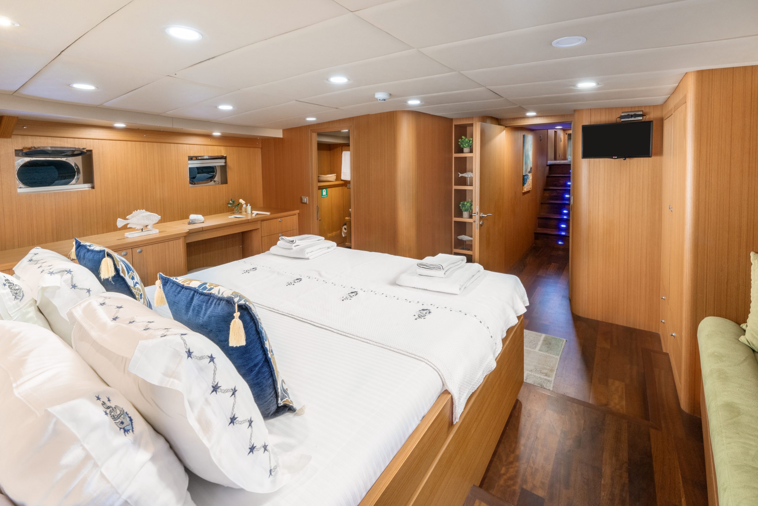 A spacious yacht cabin with a large bed, white linens, blue-and-white pillows, wooden walls, small windows, a TV, shelves with plants, and soft lighting create a cozy, modern atmosphere.