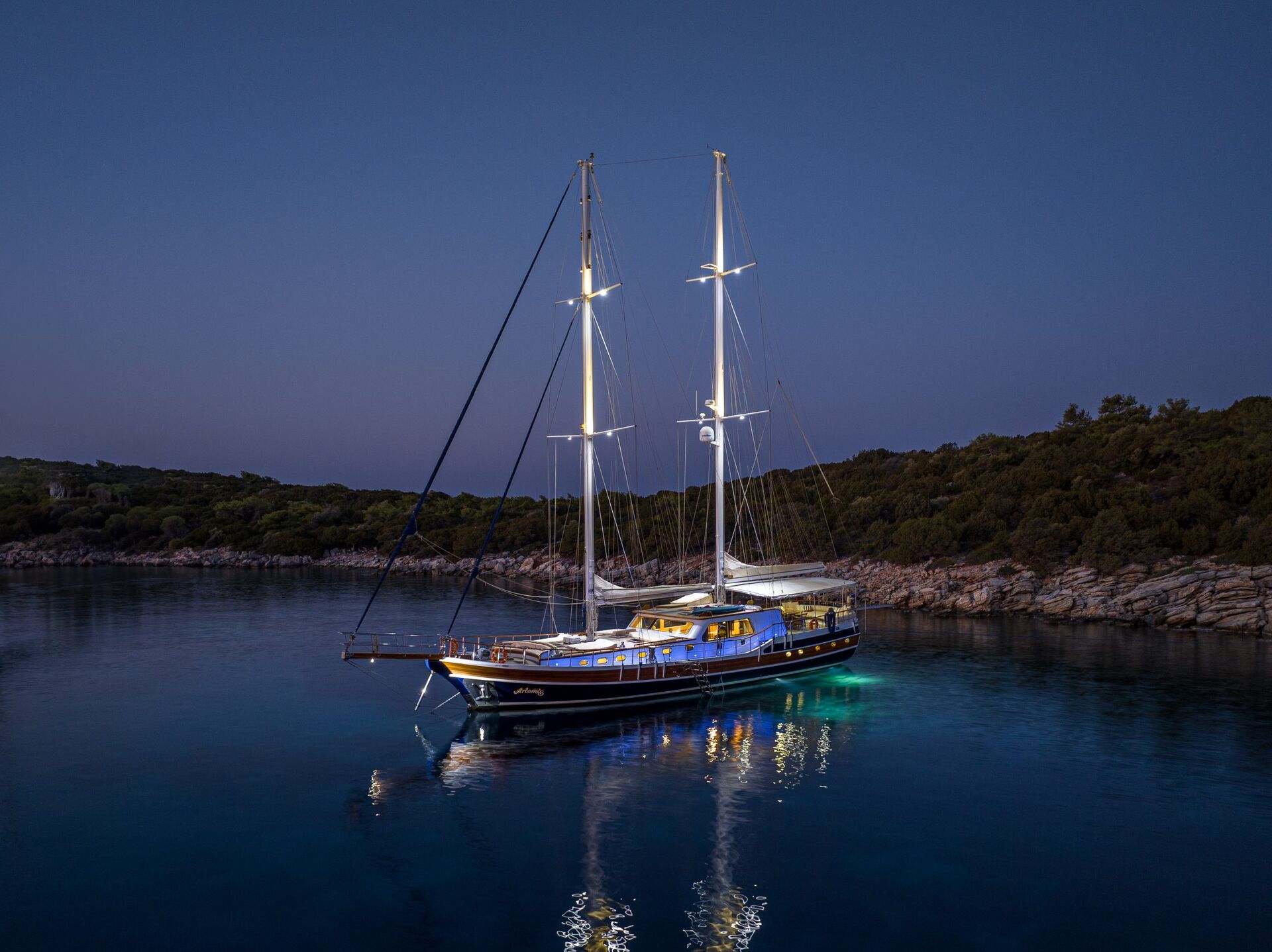 A large sailboat with illuminated deck lights is anchored in calm, clear blue water at dusk, surrounded by a tree-covered shoreline under a deep blue sky.
