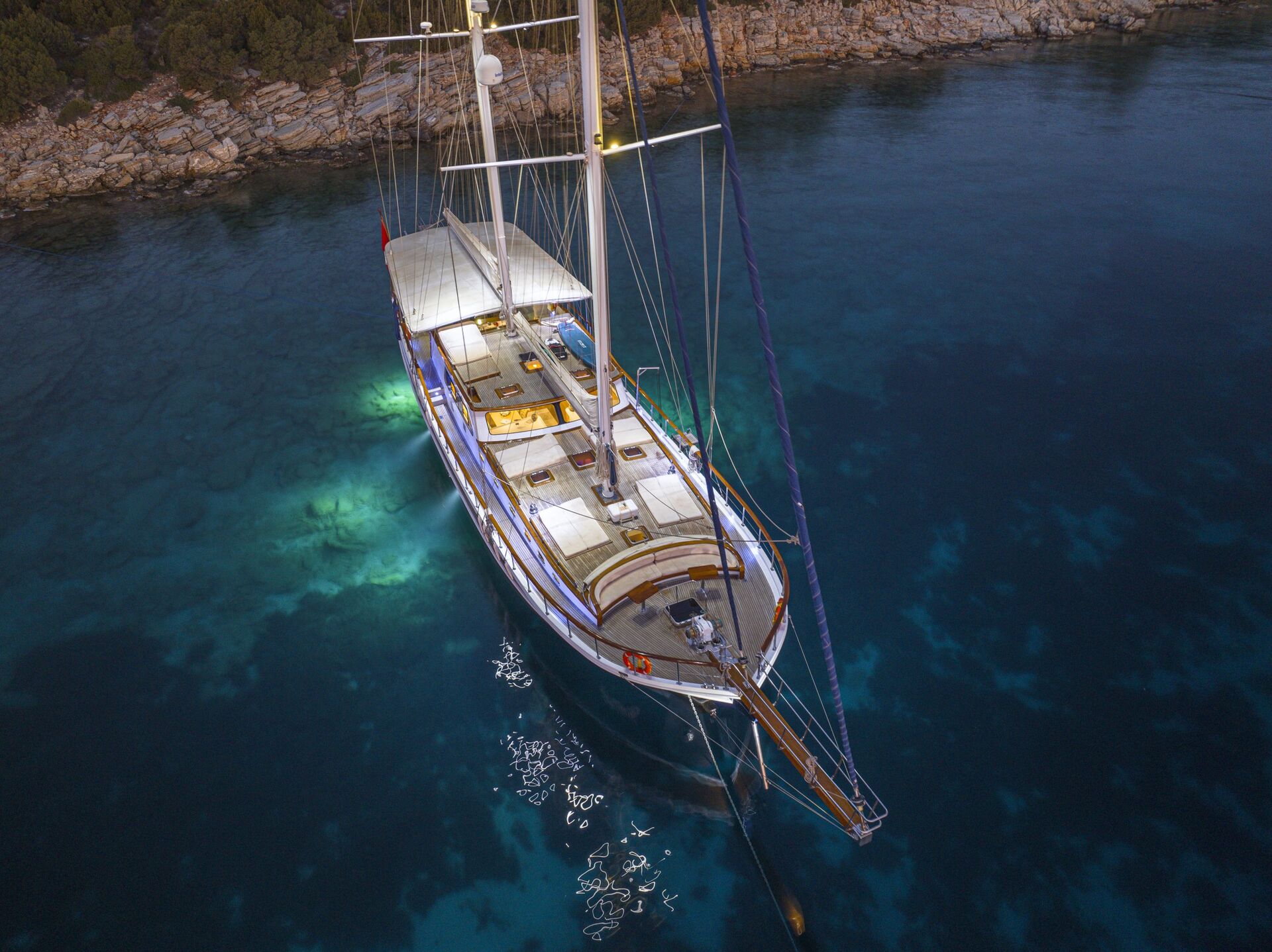 Aerial view of a large sailboat anchored in clear, calm blue water near a rocky shoreline at dusk, with underwater lights illuminating the sea around the boat.