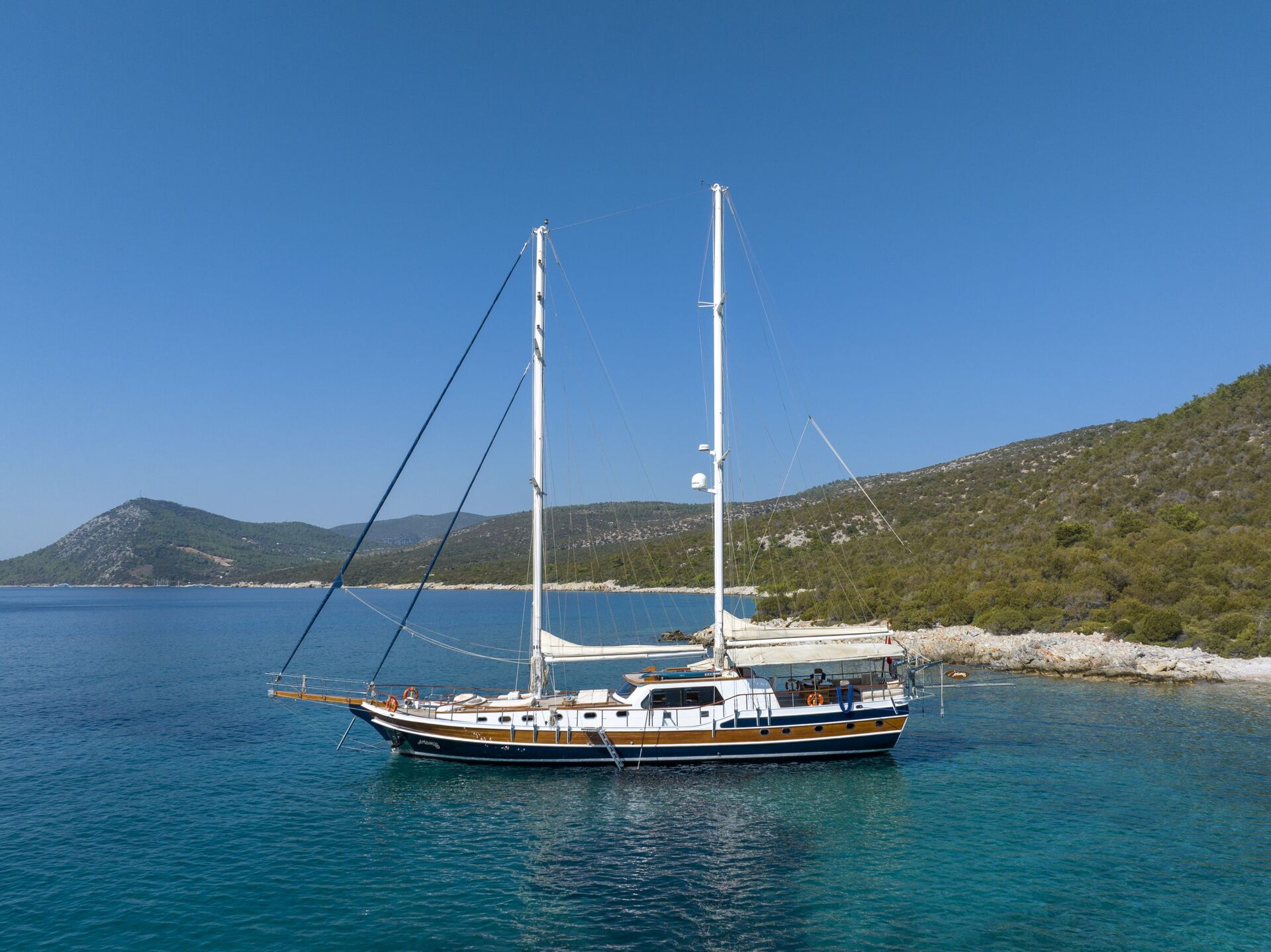 A large sailboat with two masts floats on calm, clear blue water near a rocky, tree-covered coastline. Hills rise in the background under a clear blue sky.