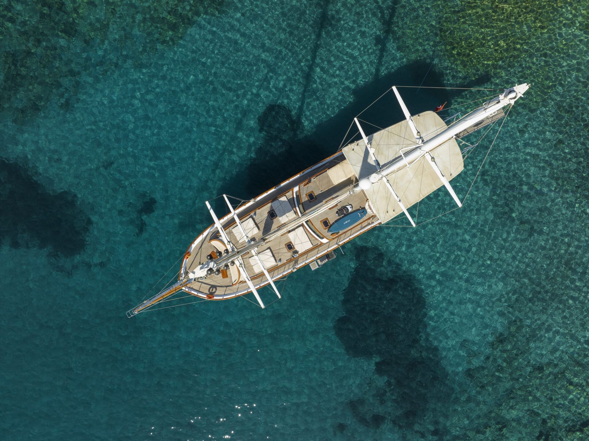 Aerial view of a sailboat anchored in clear, turquoise water with visible dark patches of underwater rocks or seaweed. The boats deck and masts are in sharp detail.