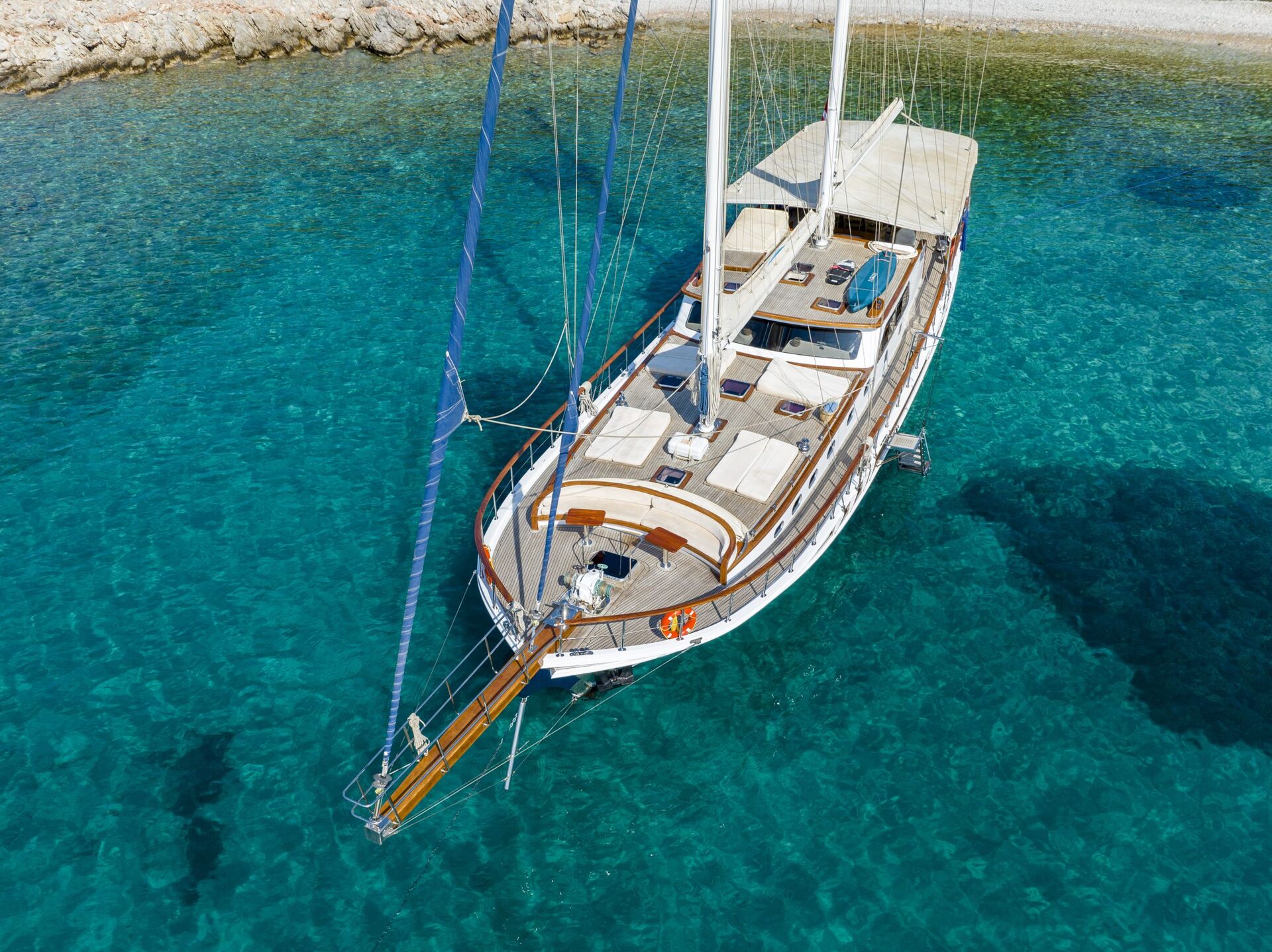 Aerial view of a large sailboat anchored in clear turquoise water near a rocky shoreline, with the boat’s decks and sails visible and sunlight highlighting the sea below.