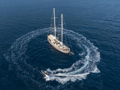 Aerial view of a large sailboat on calm blue water, with a jet ski circling around it, creating white waves in a perfect ring formation.