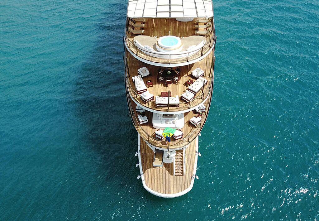 Aerial view of a luxurious yacht on clear blue water, featuring multiple wooden decks with lounge chairs, tables, and a round hot tub on the upper level. A person is sunbathing on a green towel mid-deck.