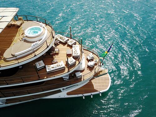 Aerial view of the deck of a luxury yacht with a hot tub, white lounge chairs, and a Brazilian flag, floating on turquoise water under bright sunlight.