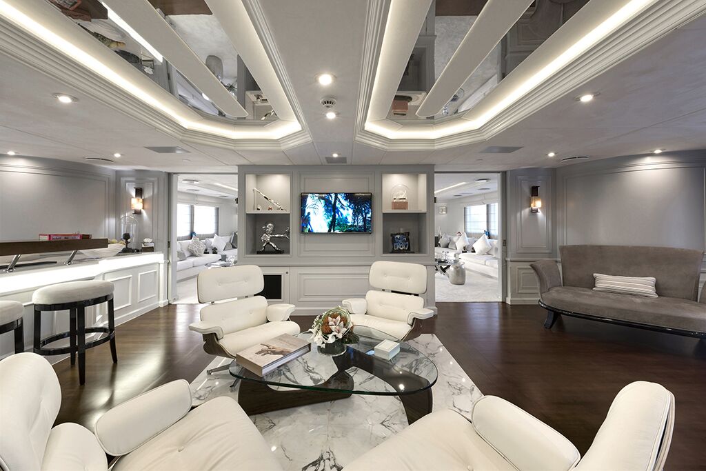 A modern, luxurious living room with white lounge chairs around a marble coffee table, a wall-mounted TV, elegant decor, and mirrored ceiling panels. The space is well-lit and features a neutral color palette.