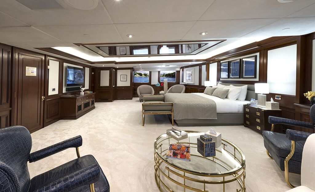 Spacious, luxurious bedroom with a large bed, plush carpet, elegant furniture, dark wood walls, two armchairs, a glass coffee table, and modern decor. Multiple windows and lights create a bright, inviting atmosphere.