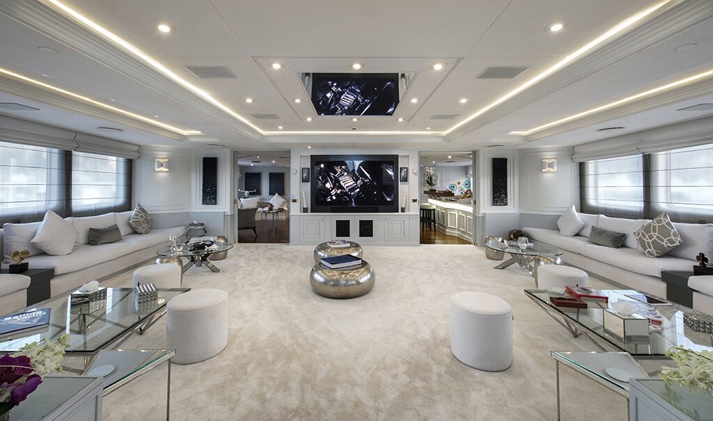 A spacious, modern living room with white sofas, glass tables, round stools, a plush beige carpet, and two large TVs on the far wall. Soft lighting and large windows brighten the room.