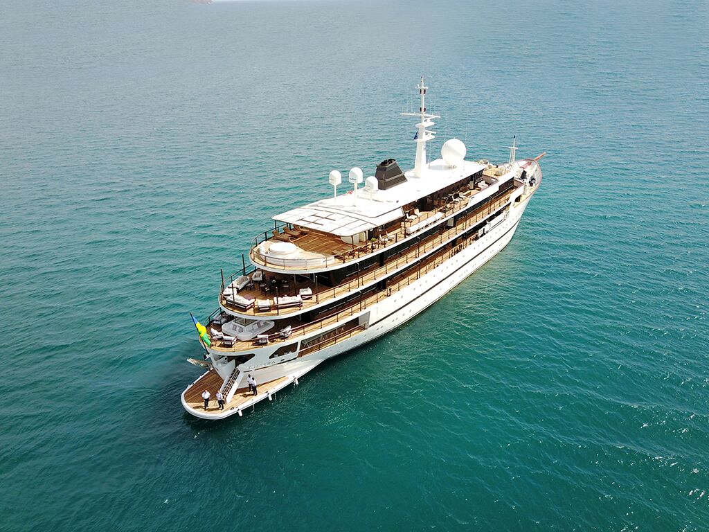 A large, luxurious white yacht with multiple decks floats on calm, turquoise ocean water. The yacht features outdoor seating areas and a flag at the stern. The scene is peaceful with no other boats nearby.