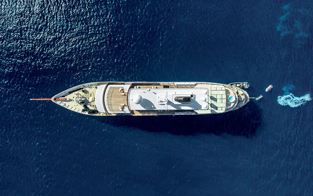 Aerial view of a large yacht anchored in deep blue water, with a small boat and a jet ski nearby, leaving a white trail in the sea.