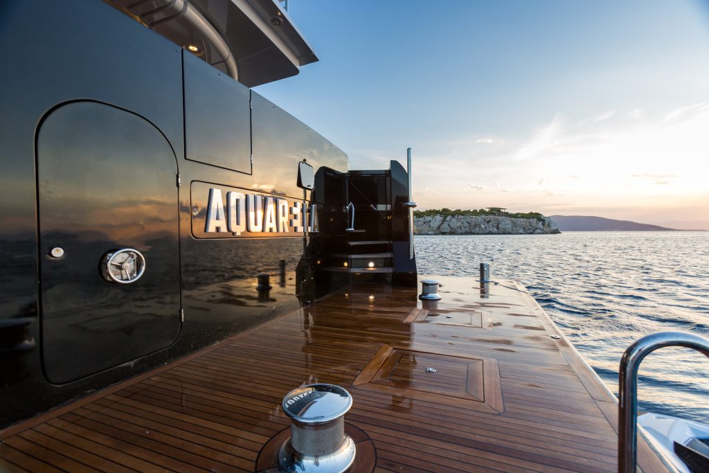 View from the deck of a luxury yacht named AQUARELLA at sunset, showing polished wood flooring, a reflective black wall, and calm sea with a rocky shoreline and distant hills.
