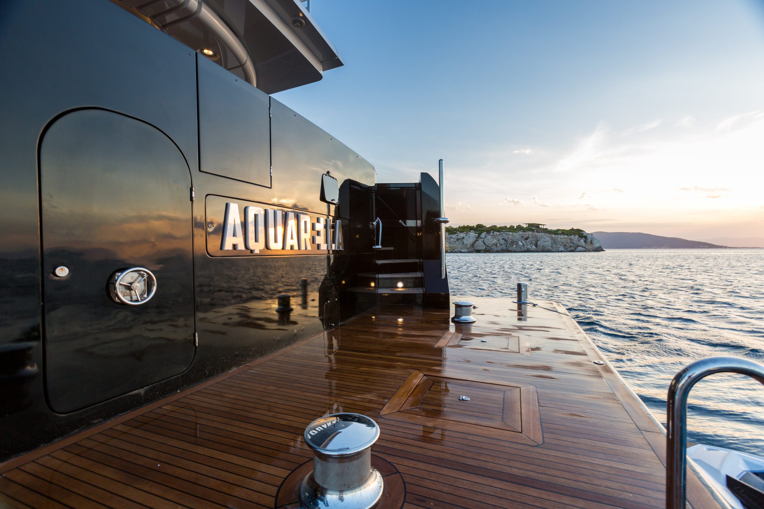 View from the deck of a luxury yacht named AQUARELLA at sunset, showing polished wood flooring, a reflective black wall, and calm sea with a rocky shoreline and distant hills.