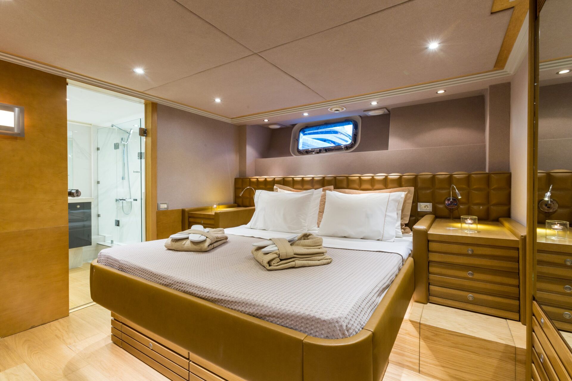 A modern yacht cabin with a neatly made double bed, folded towels, wooden furnishings, soft lighting, a small window, and an adjoining bathroom visible in the background.