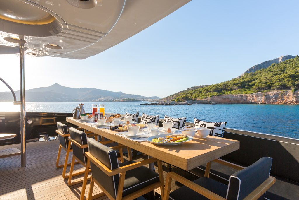 A wooden dining table set for breakfast with plates, cups, and juice sits on a yacht deck overlooking blue water and a scenic coastline with green hills under a clear sky.