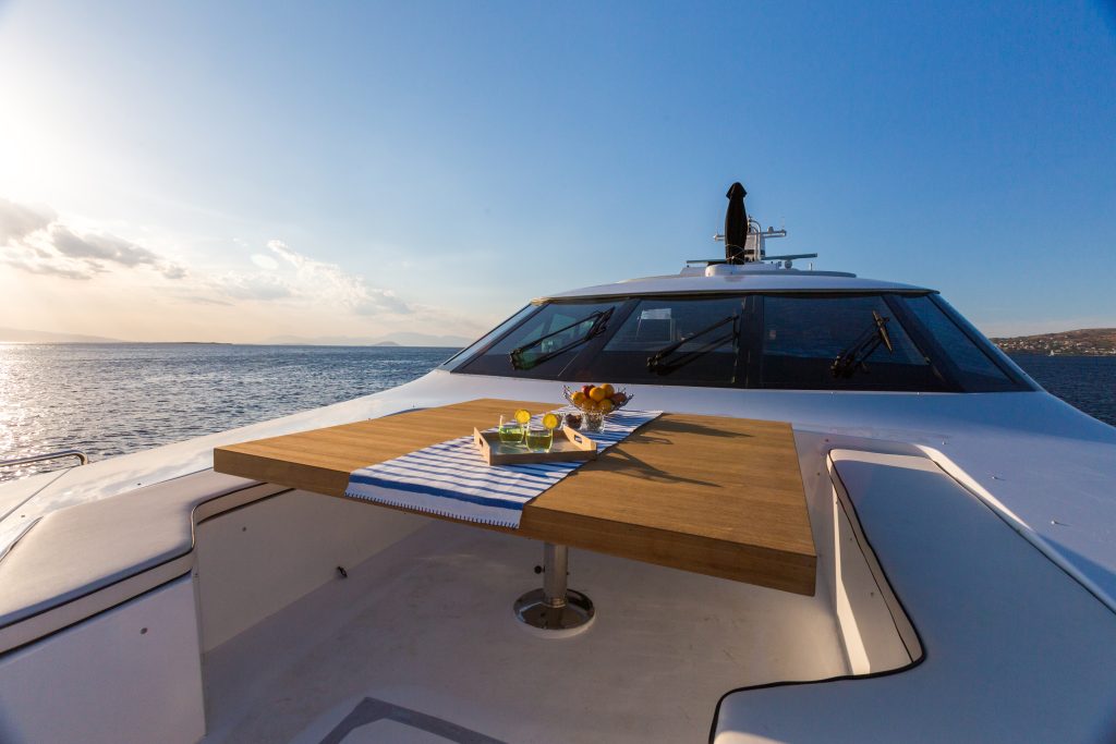 A wooden table with drinks and snacks sits on the deck of a white yacht, overlooking calm blue water and a distant shoreline under a clear sky at sunset.