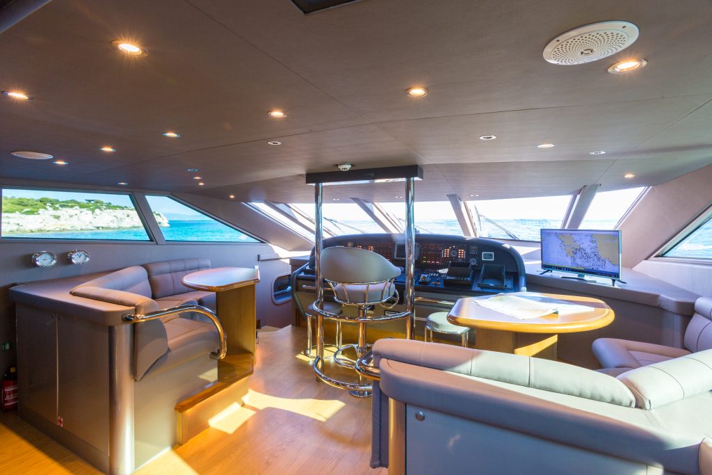 Luxurious yacht bridge with large windows, modern navigation equipment, chart table with maps, plush gray seating, and a captains chair, overlooking blue ocean and a distant coastline under bright daylight.