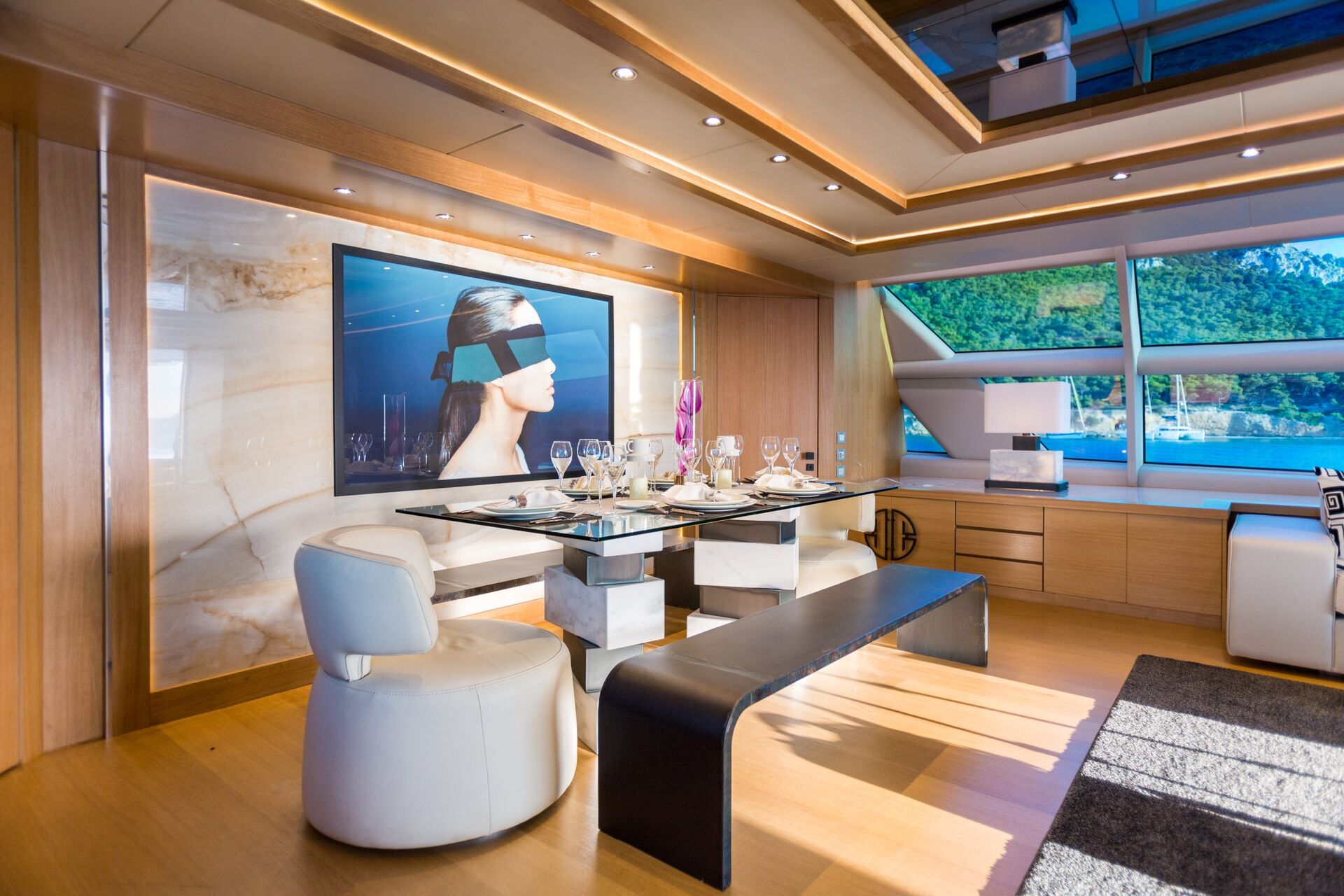 Modern yacht dining area with large windows, a sleek table set for six, white and black seating, contemporary wall art of a woman, wood paneling, and a scenic view of water and greenery outside.