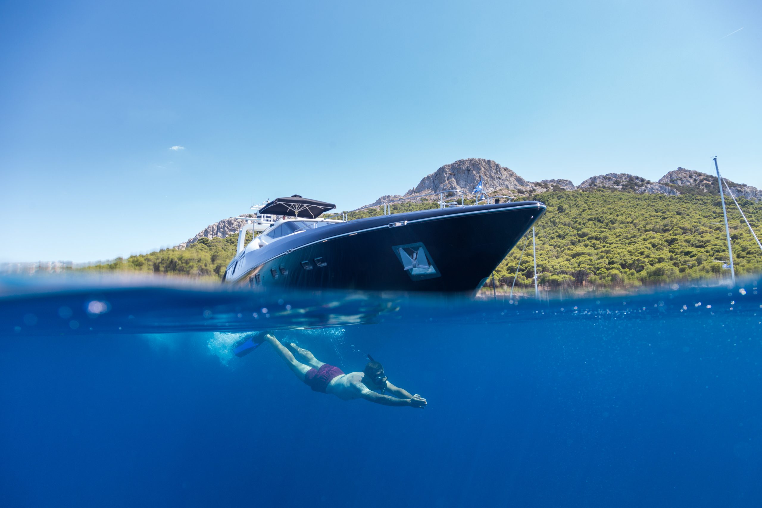 A person in swim trunks dives underwater near a large black yacht anchored in clear blue water, with a green, mountainous coastline and blue sky in the background.