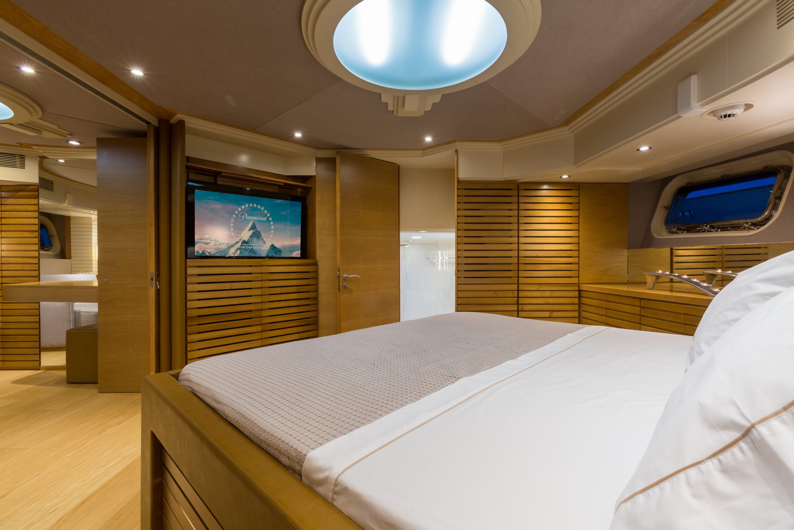 A modern yacht bedroom with wooden paneling, a large bed with white bedding, a TV showing a mountain scene, and a round ceiling light. A window and open doors reveal more interior space.