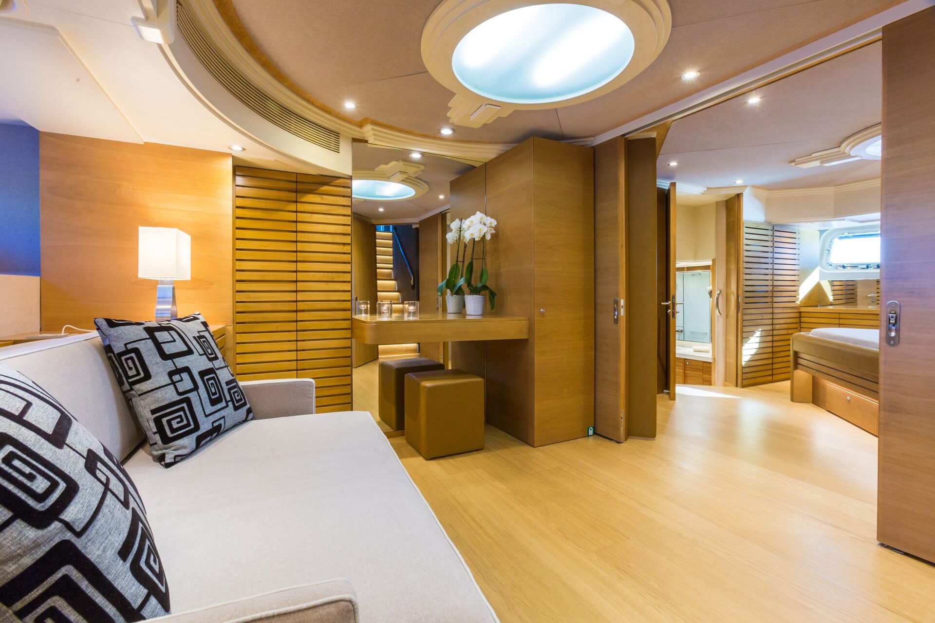 Modern yacht interior featuring light wood paneling, a beige sofa with geometric-patterned pillows, a small desk with two stools, decorative orchid, and an open doorway leading to a cozy bedroom. Natural light brightens the space.