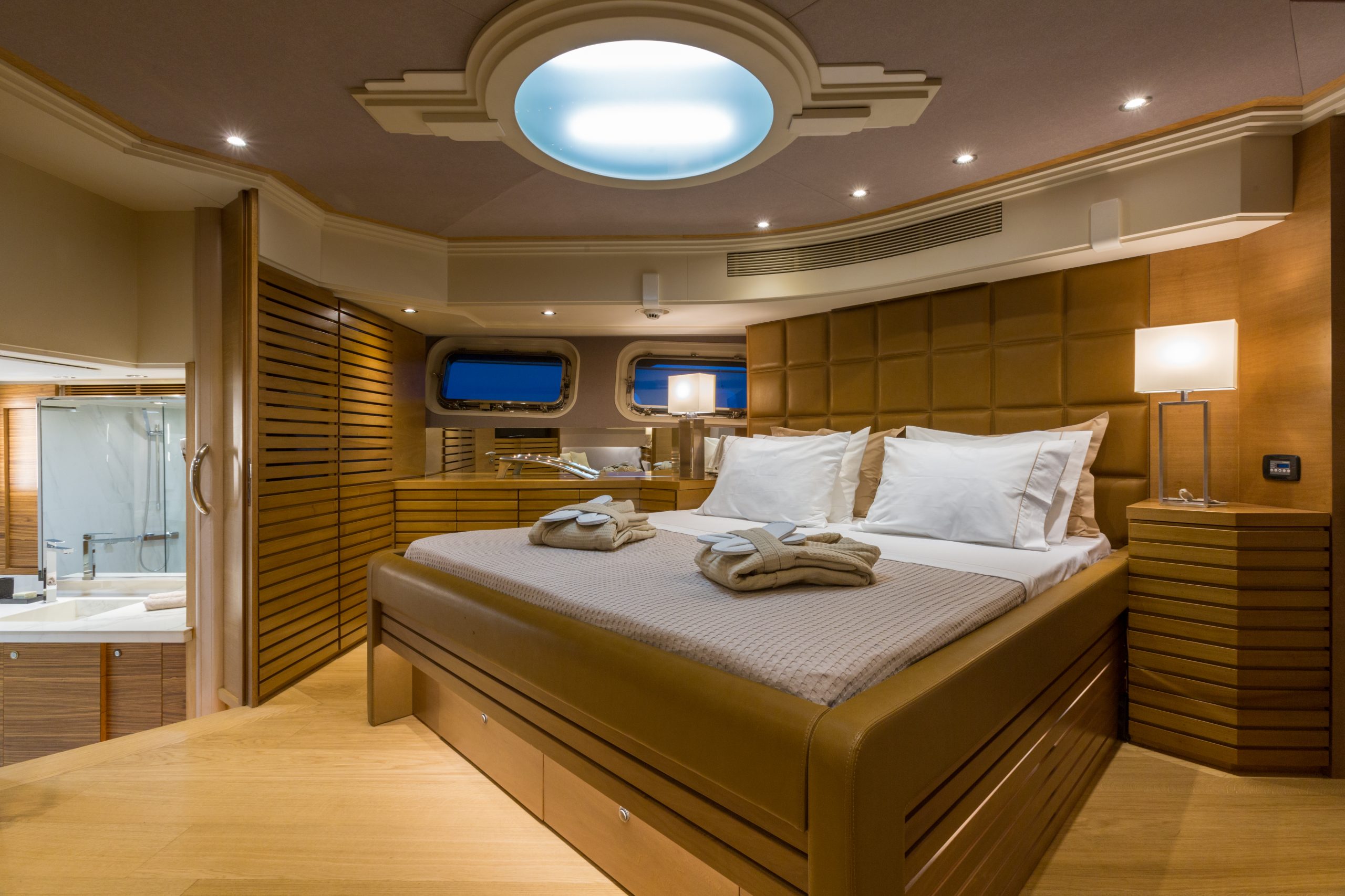 Luxurious yacht bedroom with a large bed, cushioned headboard, two robes neatly placed on the bed, warm lighting, wood-paneled walls, and small windows; a modern bathroom is visible to the left.