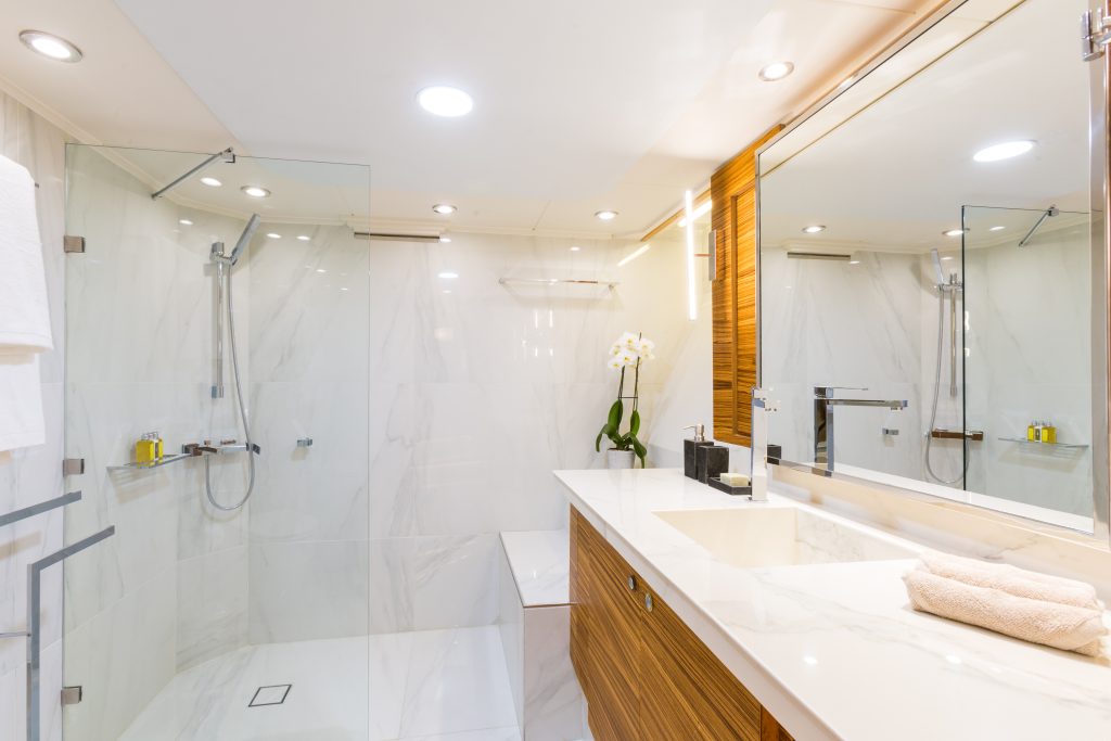 Modern bathroom with a large mirror, marble countertop, neatly folded towels, glass-enclosed shower, rain showerhead, and an orchid in a vase on the counter. Warm lighting creates a clean, inviting atmosphere.