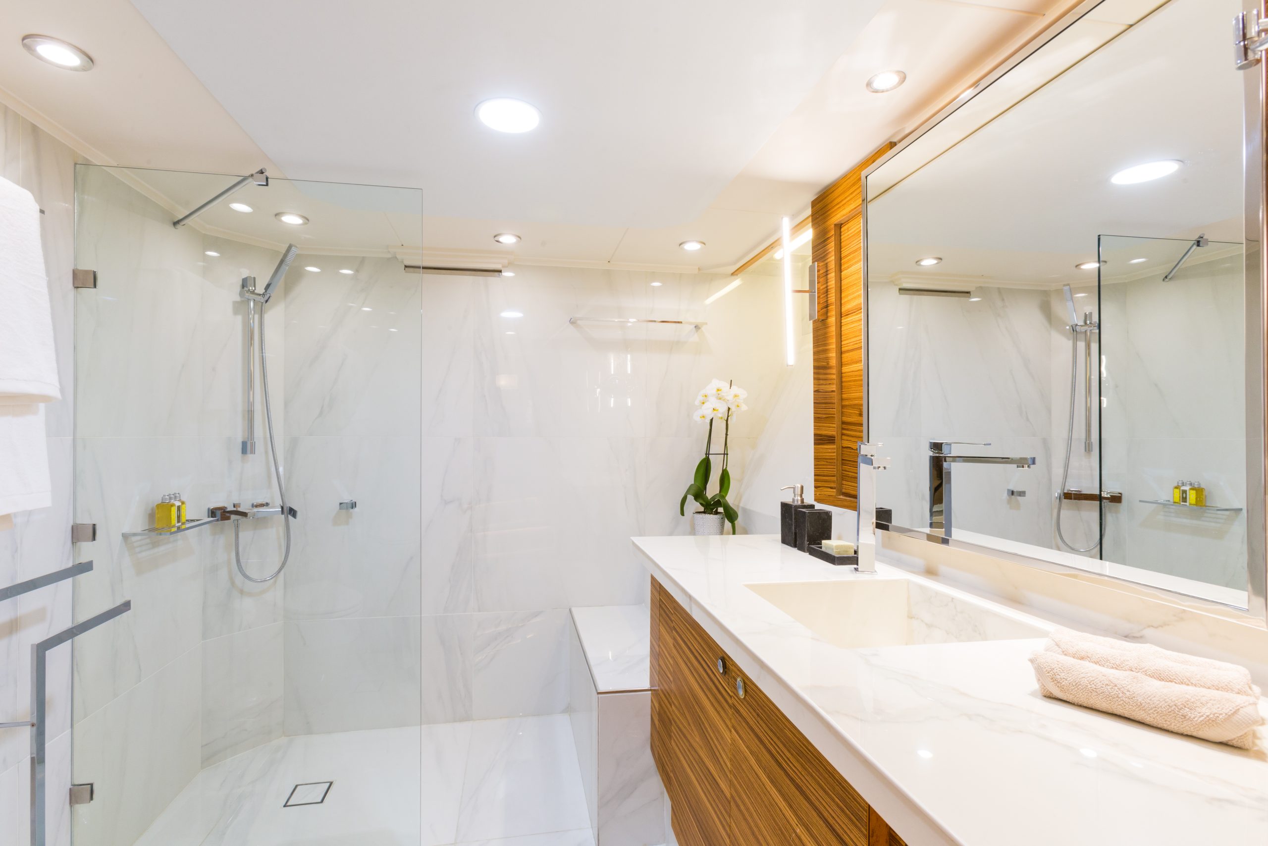 Modern bathroom with a large mirror, marble countertop, neatly folded towels, glass-enclosed shower, rain showerhead, and an orchid in a vase on the counter. Warm lighting creates a clean, inviting atmosphere.