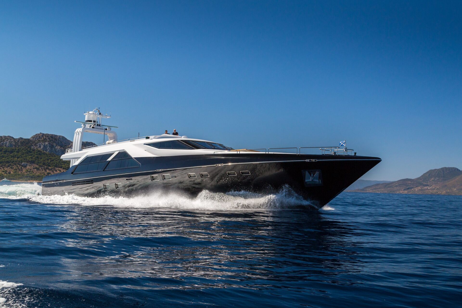 A sleek black and white luxury yacht speeds across a calm blue sea, leaving a trail of white waves, with hilly coastline and clear blue sky in the background.