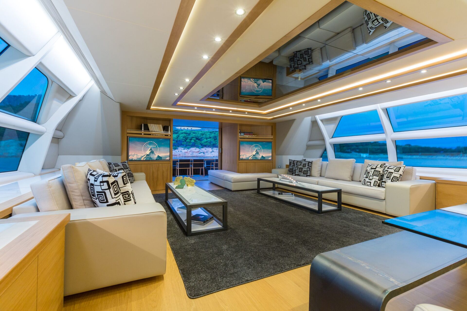 Modern yacht lounge with large windows, cream sofas with patterned pillows, glass coffee tables, a dark rug, and a well-lit ceiling with mirrors. There are scenic views of water and hills visible outside.