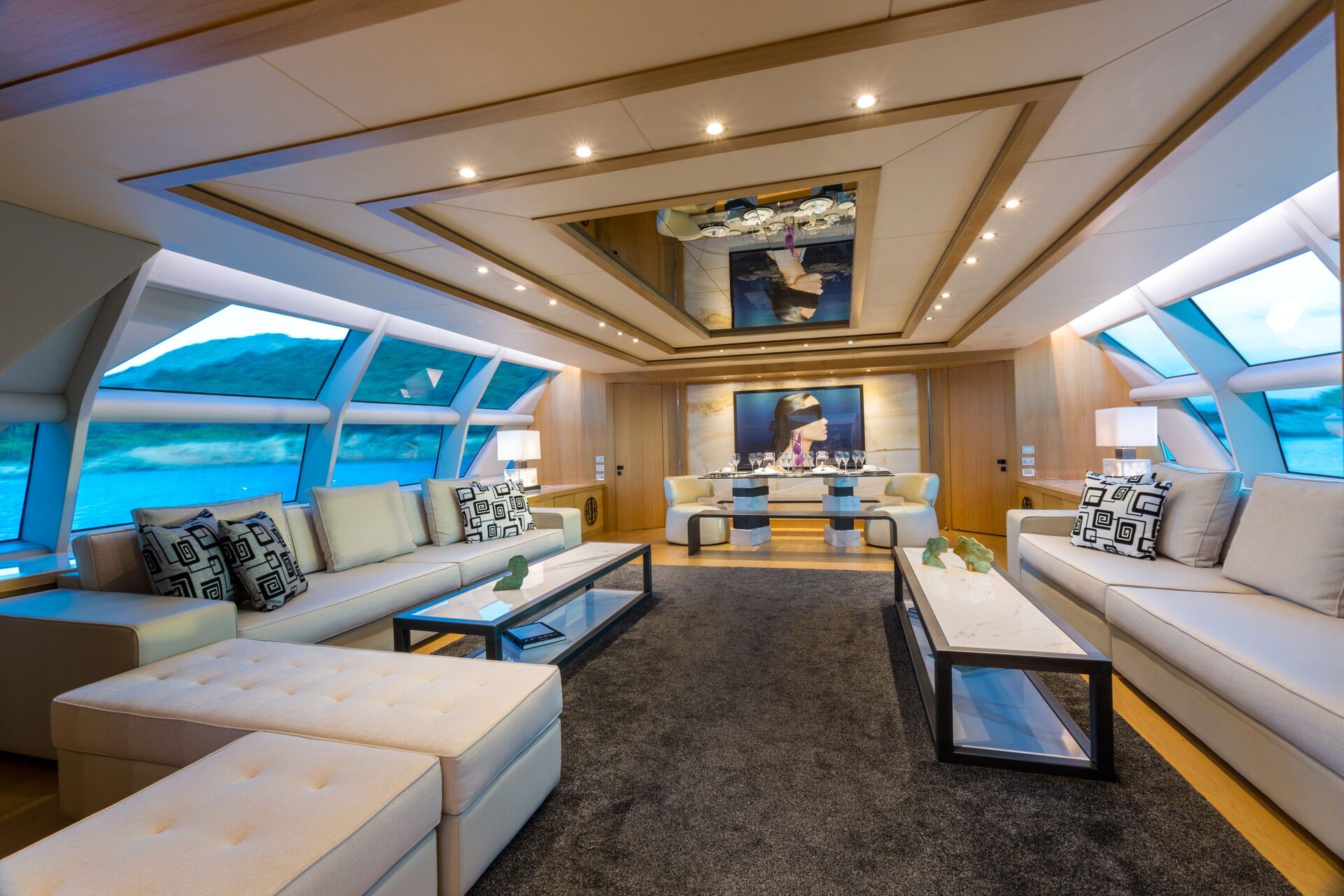 A luxurious yacht interior with large windows, beige sofas, modern glass coffee tables, decorative cushions, and artwork on the wall, all illuminated by recessed ceiling lights.