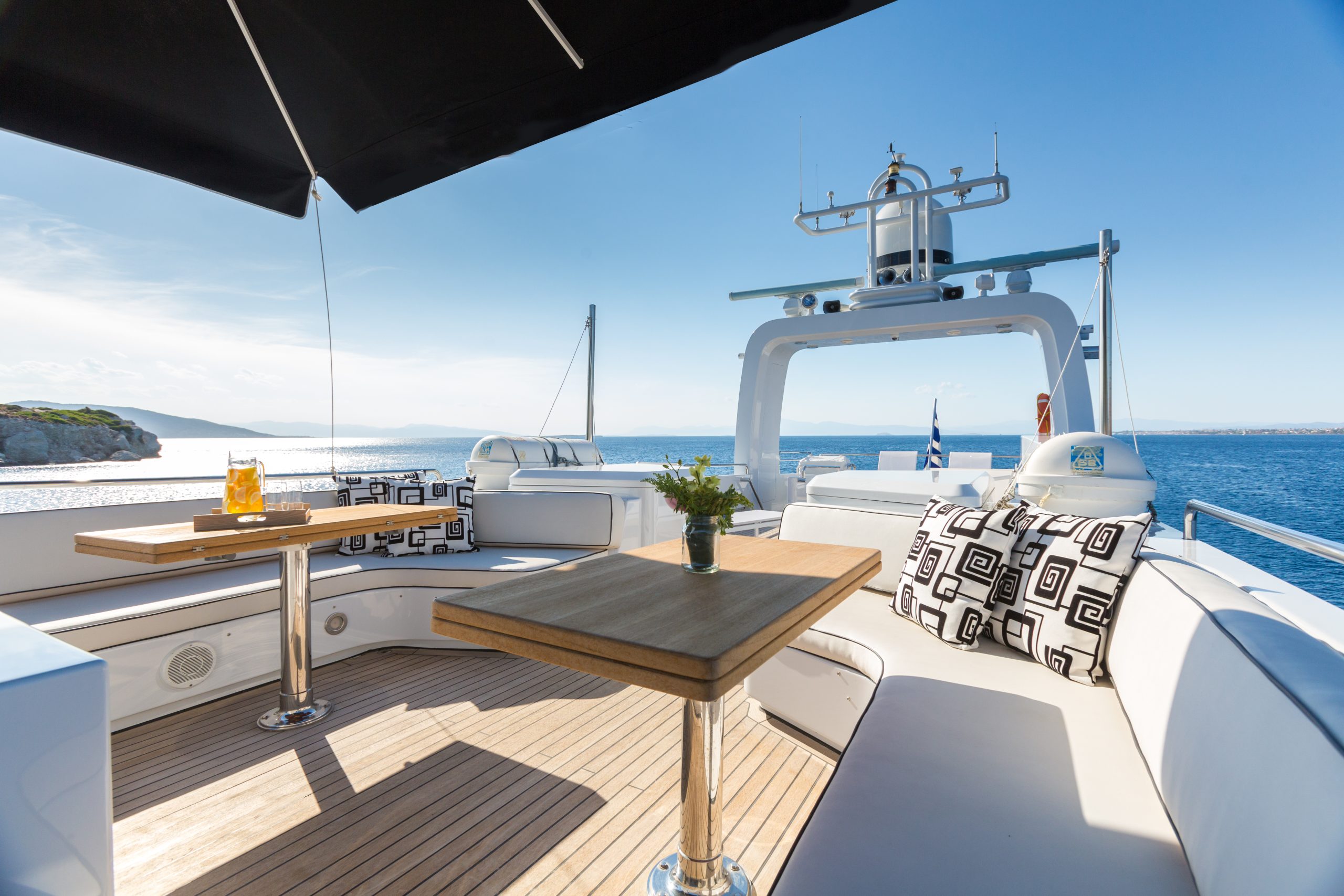 A luxury yacht deck with cushioned white seating, patterned pillows, wooden tables, drinks, and a scenic ocean view under a clear blue sky. A rocky coastline is visible in the background.