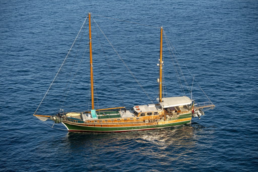 A large wooden sailboat with two masts is floating on calm blue ocean water. The boat has a covered deck area, lifeboats at the rear, and appears to be anchored or moving slowly.