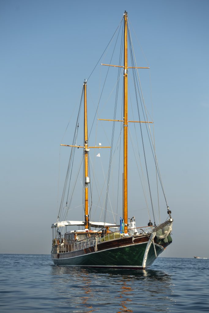 A large wooden sailboat with two tall masts floats on calm, blue water under a clear sky. The boat is viewed from the front, and there are no sails unfurled.